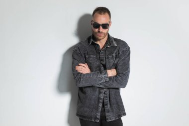 Kot ceketli havalı bir adam kollarını kavuşturup stüdyoda gri arka planda poz veriyor.