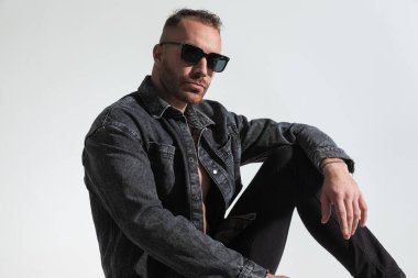 Gri kot ceketli, güneş gözlüklü, dizinde dirseğiyle poz veren, stüdyoda gri arka planda oturan sıradan bir adam.