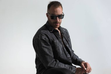 Gri kot ceket ve güneş gözlüğü takan havalı sakallı adamın portresi stüdyoda gri arka planda poz veriyor.