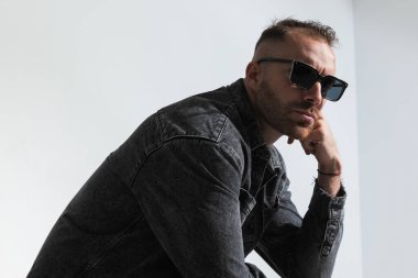 Güneş gözlüklü, el ele tutuşan ve stüdyodaki gri arka planı hayal eden sıkkın genç adamın yan görüntüsü.