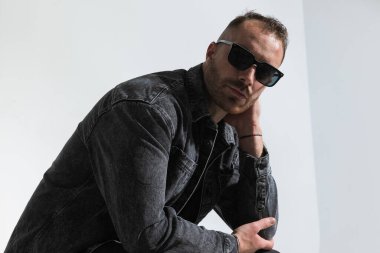 Stüdyoda gri arka planda otururken el ele tutuşan ve dirseğiyle dizlerinin üzerinde poz veren çekici genç adamın yan görüntüsü