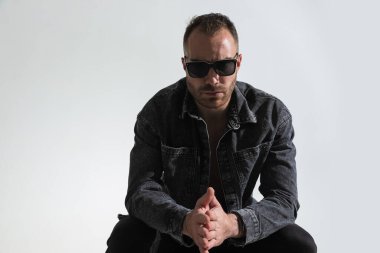 Güneş gözlüğü takan, kot ceket giyen, avuçlarını ovuşturan, gri arka planda oturan kendine güvenen bir adam.