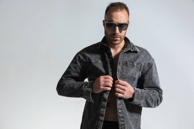 Güneş gözlüklü, gri kot ceketli, kendine güvenen sakallı bir adamın portresi. Düğmelerini ilikliyor ve stüdyoda gri arka planda poz veriyor.