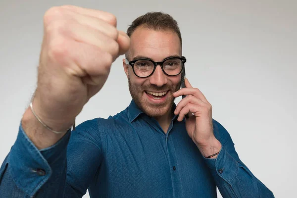 年轻而随意的男人正在庆祝他通过电话得到的好消息 穿着蓝色衬衫 戴着眼镜 背景是灰色的工作室 — 图库照片