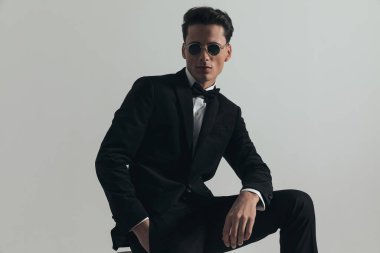 Siyah smokinli seksi genç damat güneş gözlüğü takıyor ve el ele tutuşurken stüdyoda gri arka planda çömeliyor.