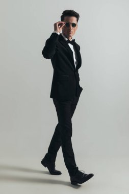 Stüdyoda gri arka planda yürürken siyah smokin giyen ve güneş gözlüğü takan şık genç bir damadın tam vücut resmi.