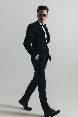 Siyah smokin ve güneş gözlüğü takan, cebinde el ele tutuşan ve stüdyoda gri arka planda yürüyen esmer saçlı çekici modern adamın yan görüntüsü.