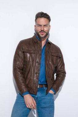 Seksi ve şehvetli bir adam bir elini cebine sokuyor, kahverengi deri ceket giyiyor, beyaz stüdyo arka planında gözlük takıyor.