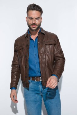 Beyaz stüdyo geçmişine karşı poz veren, kahverengi deri ceketli ve gözlüklü seksi bir adam.