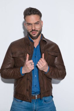Genç bir adam ceketini çekiyor ve beyaz stüdyo geçmişine karşı gözlük takıyor.
