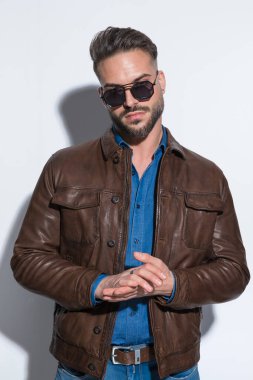 Çekici sıradan bir adam avuçlarını ovuyor, güneş gözlüğü takıyor ve beyaz stüdyo arka planına karşı kahverengi deri ceket giyiyor.