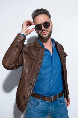 Erkeksi çerçeveli sıradan bir adam güneş gözlüklerini ayarlıyor ve beyaz stüdyo arka planına karşı kahverengi deri ceket giyiyor.