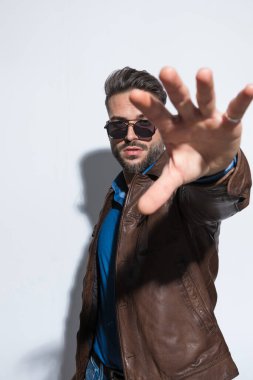 Genç bir adam çerçeveyi tek eliyle gölgede bırakıyor ve güneş gözlüğü takıyor, kahverengi deri ceket giyiyor.