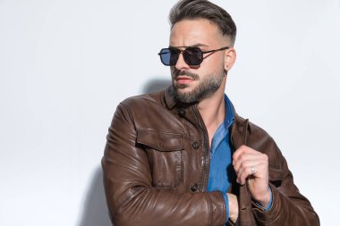 Genç adam bir elini ceketinin cebine sokuyor, güneş gözlüğü takıyor ve beyaz stüdyo geçmişine bakıyor.