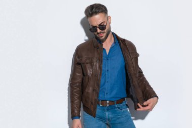 Dramatik sıradan bir adam güneş gözlüğü ve kahverengi deri bir ceketle beyaz stüdyo arka planına bakıyor.