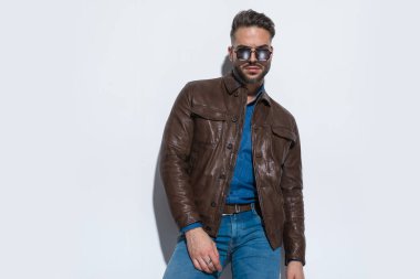Beyaz stüdyo arka planında kahverengi deri ceket giyen ve güneş gözlüğü takan seksi bir adamla moda fotoğrafı.