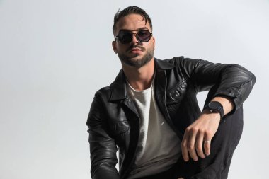 Güneş gözlüklü yakışıklı adam diziyle poz veriyor ve stüdyoda gri arka planda otururken kendine güveniyor.