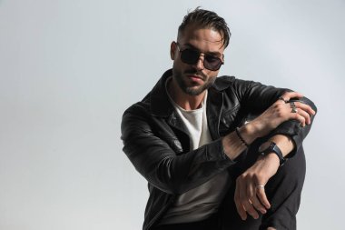 Modern saç stili, şık deri ceket ve güneş gözlüğü takan, gri arka planda dirseğiyle poz veren, kendine güvenen havalı bir adam.