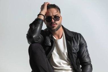 Kendine güvenen sakallı bir adamın portresi. Deri ceket ve güneş gözlüğü takıyor. Stüdyonun gri arka planında dirsekleriyle poz veriyor.