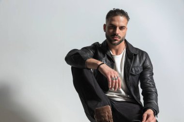 Siyah deri ceketli şık bir genç adam. Stüdyoda gri bir arkaplan önünde dirseğiyle poz veriyor.
