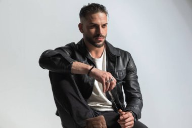 Çekici moda adamı siyah deri ceket giyiyor ve güneş gözlüğü tutarken dirseğiyle poz veriyor ve stüdyoda gri arkaplanın önüne bakıyor.