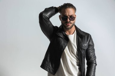 Moda esmer adam ıslak modern saç stili ayarlıyor ve stüdyoda gri arka plan önünde siyah deri ceket ve güneş gözlüğü takıyor. 