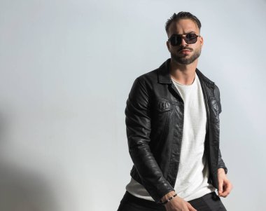 Siyah deri ceket ve güneş gözlüğü takan seksi Latin adam gri arka planda havalı bir şekilde poz veriyor. 