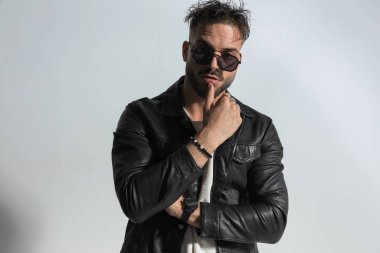 Modern saç stili çapraz kollu, tıraş edilmemiş bir adamın portresi. Stüdyoda gri arka planda dudaklarına dokunuyor.