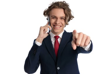 Takım elbiseli, kıvırcık saçlı mutlu adam telefonda konuşuyor, işaret ediyor ve stüdyoda beyaz arkaplan önünde seçim yapıyor.