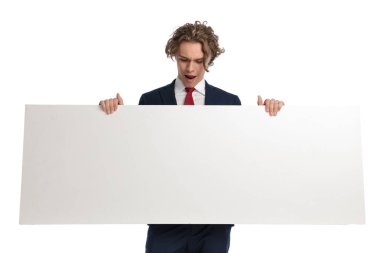 Genç iş adamının elinde yatay olarak boş bir tahta tutarak stüdyoda beyaz arka planda şok olmuş bir yüz ifadesi oluşturmasına şaşırdı.