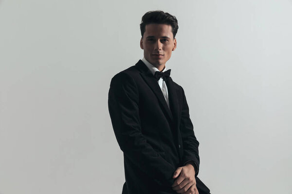 portrait of happy young groom wearing black tuxedo with bowtie, holding hand together and smiling in front of grey background