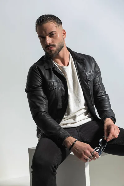 fashion bearded man in leather jacket with wet hair holding sunglasses and sitting in a fashion pose in front of grey background in studio, portrait