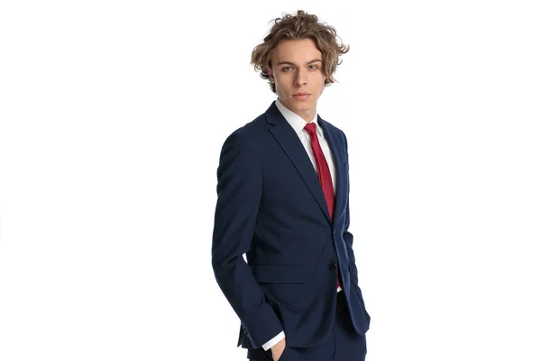 スタジオの白い背景の前に立っている間ポケットに手でポーズをとった赤いネクタイでエレガントなスーツを着た魅力的な若いビジネスマンの肖像画 — ストック写真