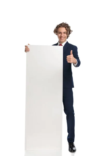 スーツ姿のハンサムなビジネスマン白い空の板と笑顔を提示しながら親指を作りますスタジオで白い背景の前でジェスチャー — ストック写真