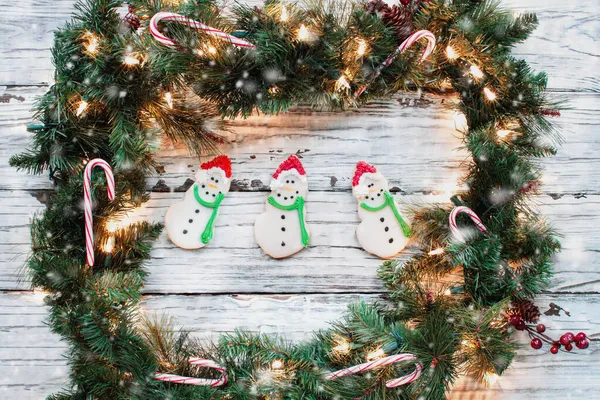 白い素朴な木の背景に3つのアイスクリスマススノーマンクッキーでクリスマスガーランドで作られた休日の花輪 フラットレイまたはトップビュー位置から撮影された画像 — ストック写真