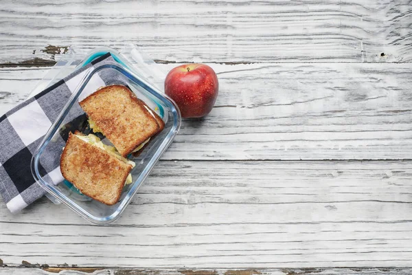 Packed lunch of fresh apple and home egg salad sandwich with toasted wheat bread in a glass container. Healthy vegetarian diet concept over a rustic white wooden background. Table top view.