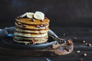 Pancakes served with fresh banana fruit and nuts with maple syrup dripping down the sides. Selective focus with blurred background and foreground.
