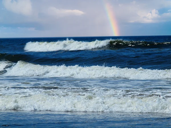 Onde oceaniche che si infrangono sulla riva con un arcobaleno parziale sullo sfondo — Foto Stock