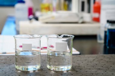 Two beakers with clear liquids and blank labels on a pad in a laboratory