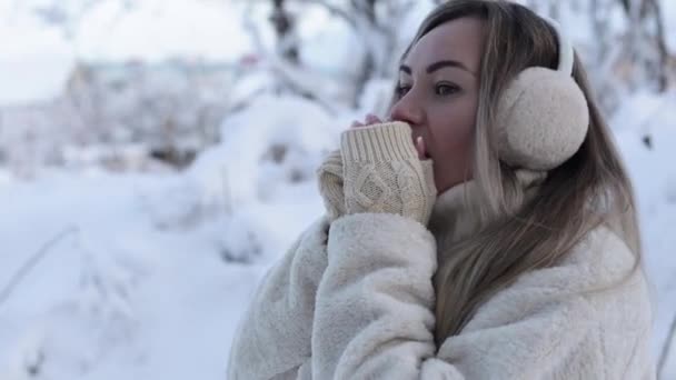 Portrait of a cute girl in winter in warm headphones and knitted mittens. The girl warms her hands breathing on them with her mouth on the street. Winter cold — Vídeo de Stock