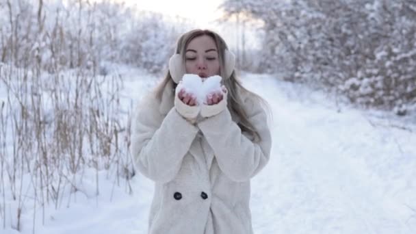 A beautiful girl in a winter coat and headphones blows snow from the gloves of a snowball in the form of a heart.Winter pampering in the forest with snow — Vídeo de Stock