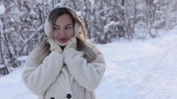 Portrait of a cute girl in winter in warm headphones and knitted mittens. The girl warms her hands breathing on them with her mouth on the street. Winter cold — Stockvideo