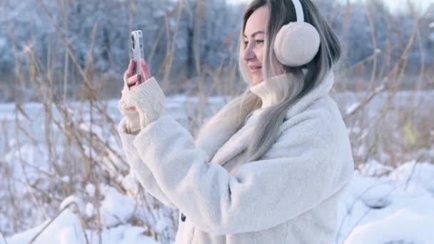 UKRAINE, KIEV - DECEMBER 27, 2021: A woman in a snowy forest in winter shoots a video on a new iPhone 13 camera. A beautiful girl walks on the street in frost — стокове відео
