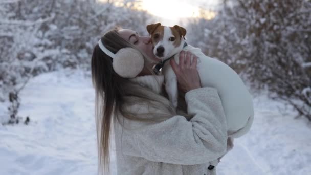 รักสัตว์ สาวน่ารักในเสื้อโค้ทสีขาวเล่นและจูบสุนัข Jack Russell Terrier ด้านนอกในฤดูหนาวในหิมะ — วีดีโอสต็อก