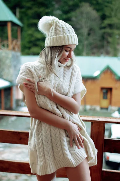 Uma menina sexy em um suéter branco e meias na varanda admira a beleza da natureza. roupas de lã e um chapéu em uma bela loira — Fotografia de Stock