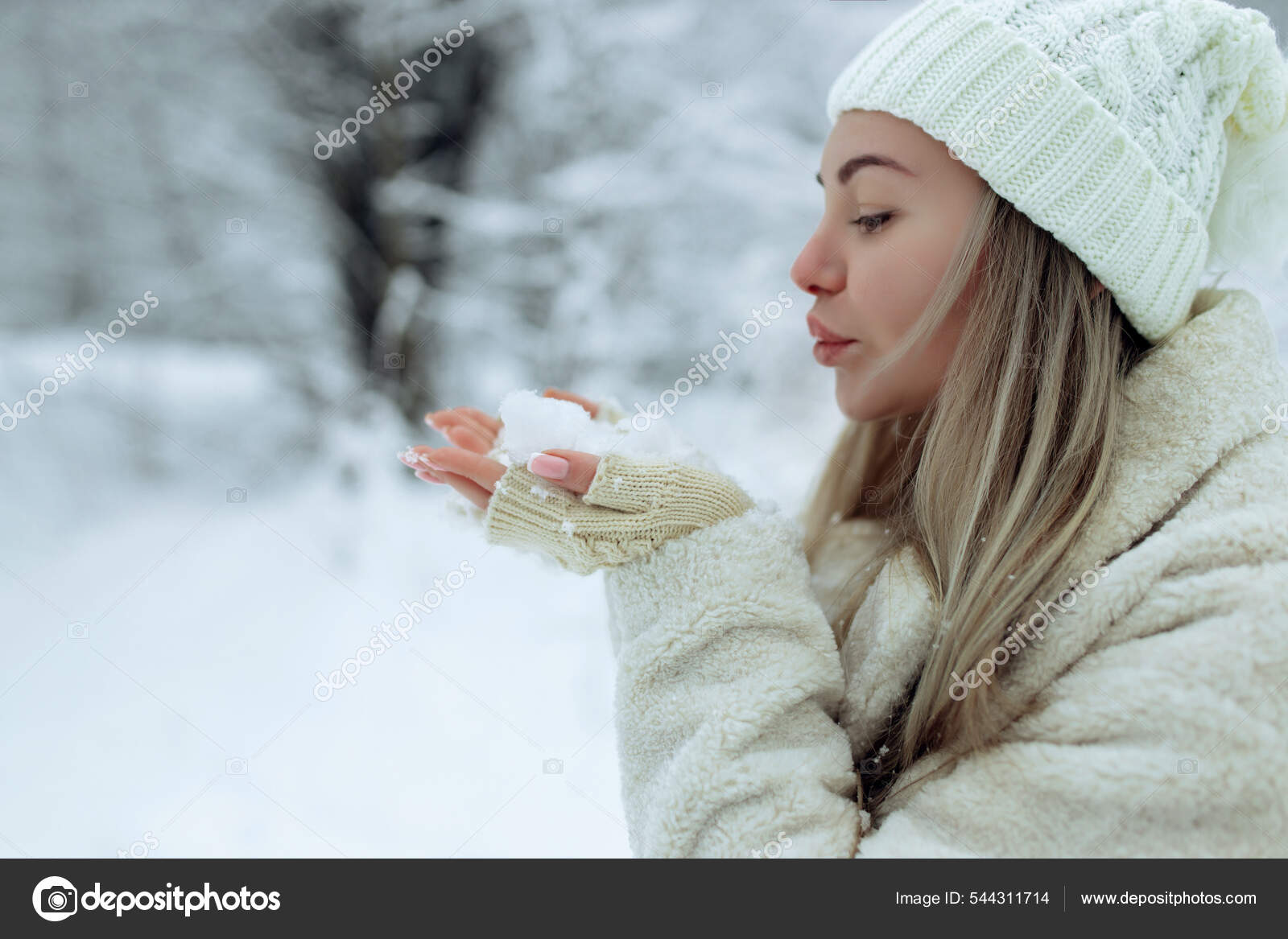 Beautiful girl in fashionable winter clothes blows snow from the palms of  her hands. Joy to the first snow .Young blonde in a white hat in a snowy  forest Stock Photo by ©