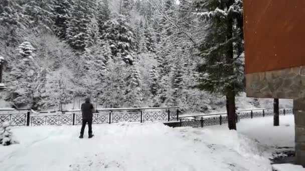 Fotógrafo - Un turista toma fotos de un bosque de pinos cubierto de nieve y montañas. Plano general de un hombre de espaldas con una cámara cerca de un bosque de pinos — Vídeo de stock