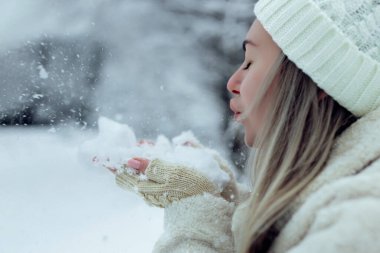 Şık kış kıyafetleri içinde güzel bir kız avuçlarından kar üflüyor. İlk kara neşe. Karlı ormanda beyaz şapkalı genç sarışın.