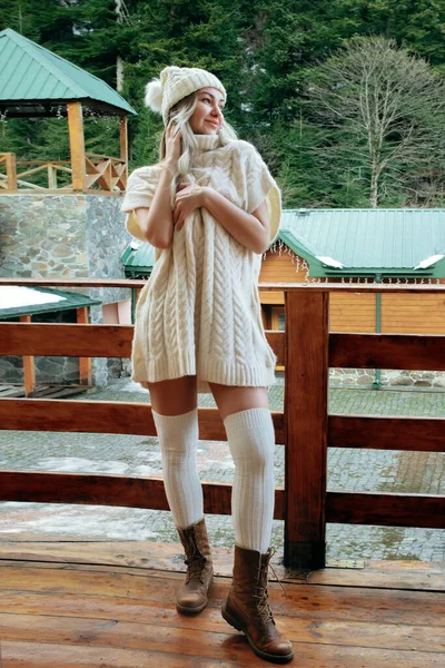 Uma menina sexy em um suéter branco e meias na varanda admira a beleza da natureza. roupas de lã e um chapéu em uma bela loira — Fotografia de Stock