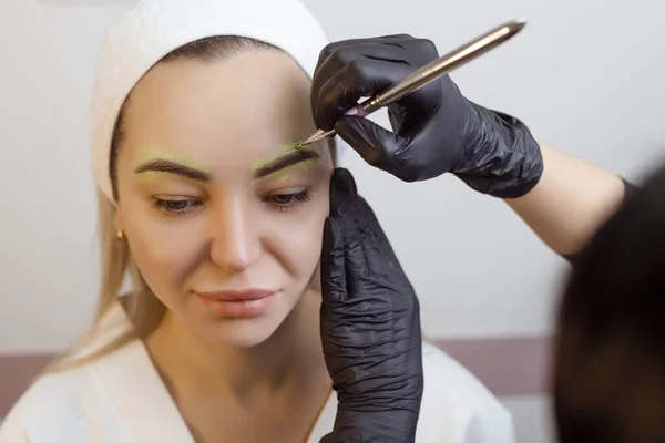 The hand of a make-up artist in black gloves applies a yellow eyebrow paste to the eyebrow. Professional stylish permanent makeup, eyebrow tinting. Close up.